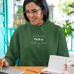 Forest green hoodie with the words Birdsong feeds my should above and below a white graphic of 4 birds on a branch. Worn by a woman working at a computer.