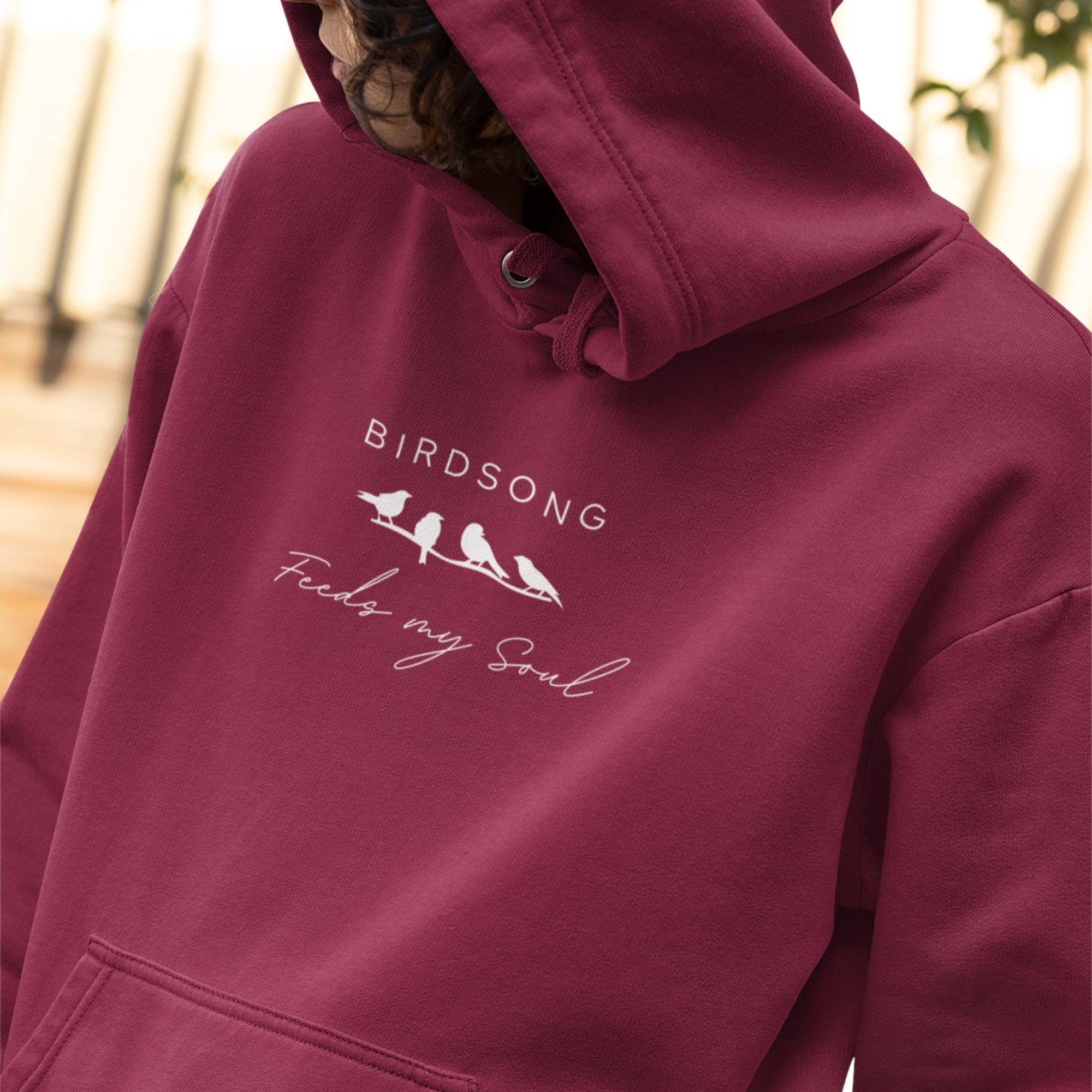Maroon hoodie with the words Birdsong feeds my should above and below a white graphic of 4 birds on a branch. Worn by a woman.