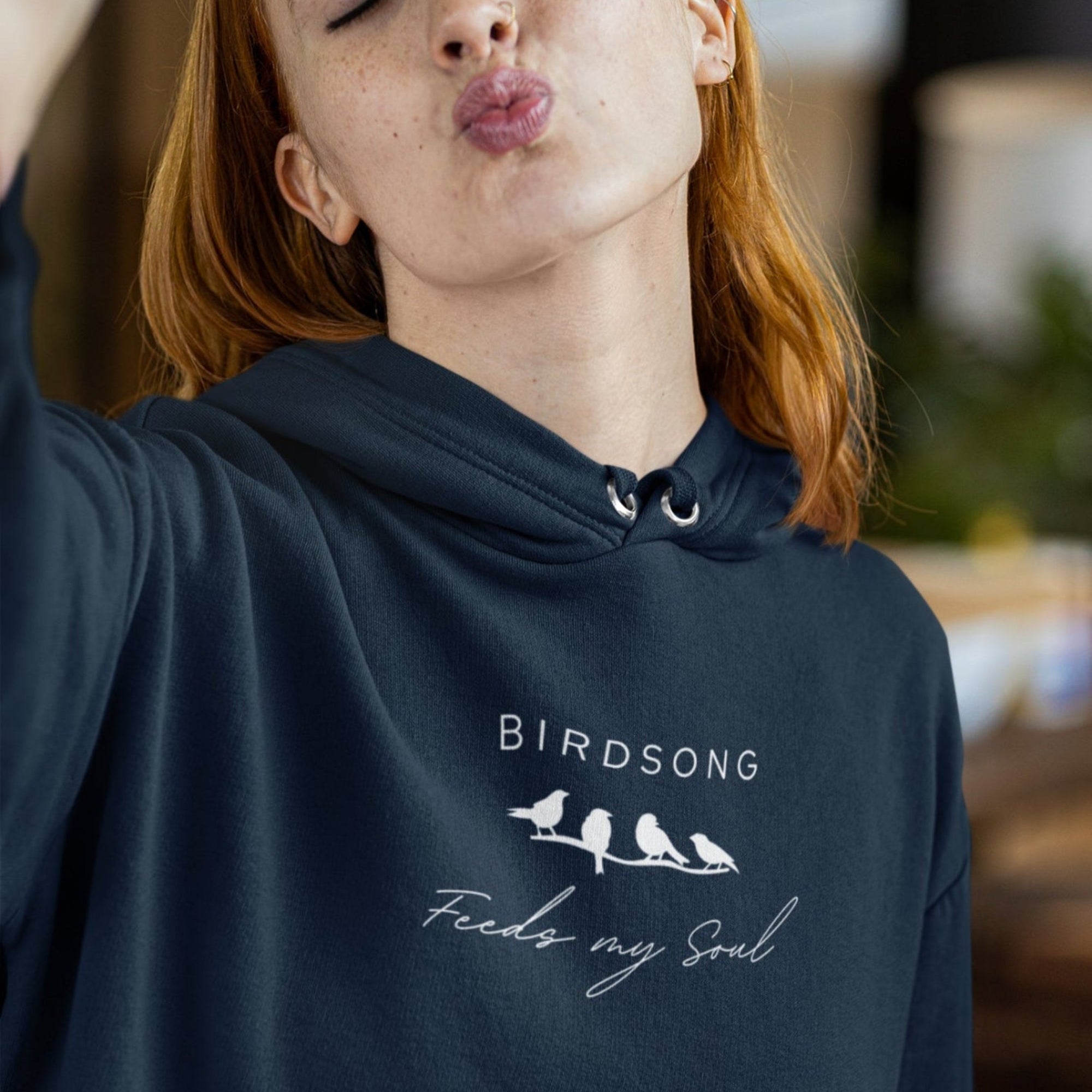 Navy hoodie with the words Birdsong feeds my should above and below a white graphic of 4 birds on a branch. Worn by a woman making a kissy face.