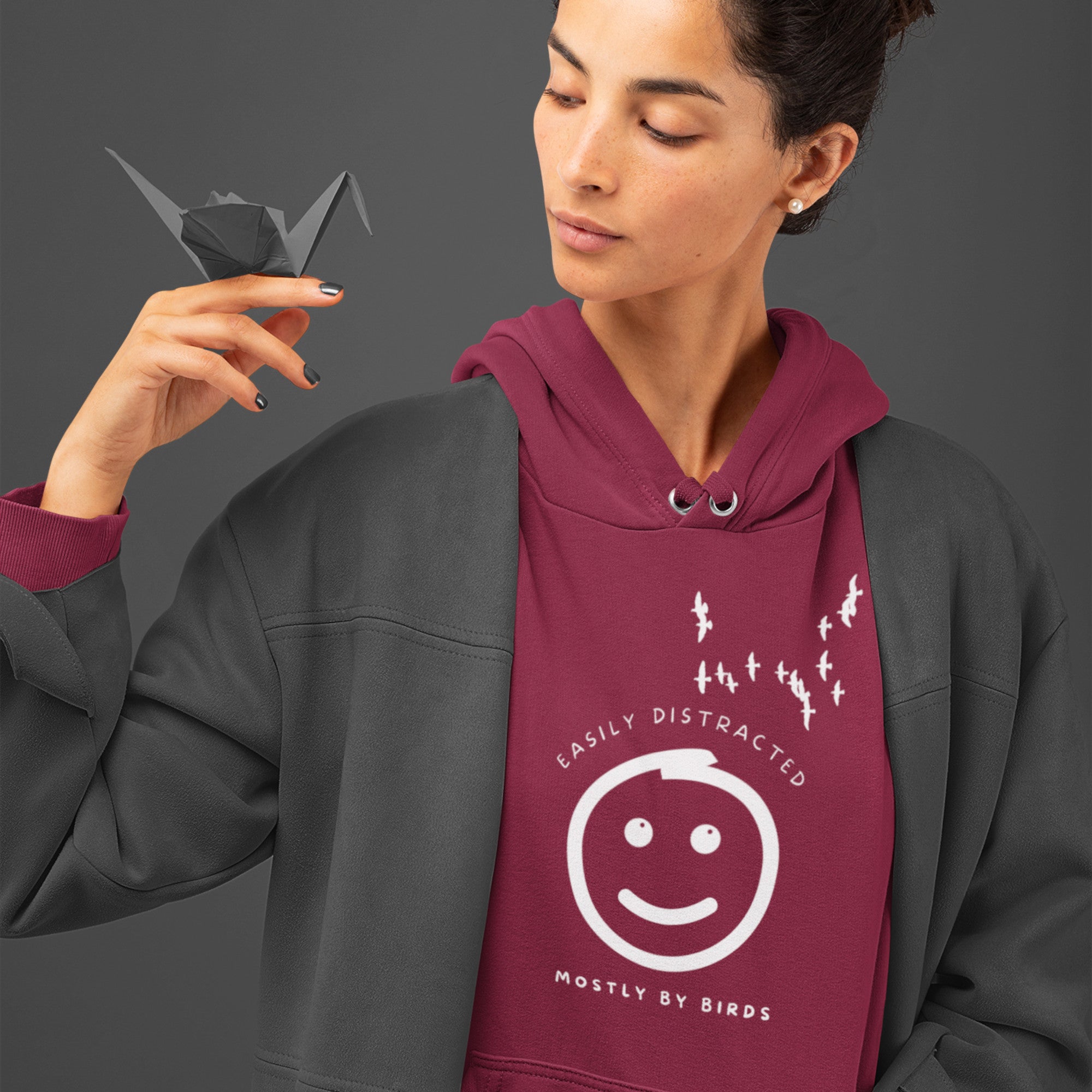 Maroon hoodie with a smiley face looking off to the right at a flock of birds and the words Easily Distracted, Mostly By Birds on the front. Worn my a woman holding an origami bird.
