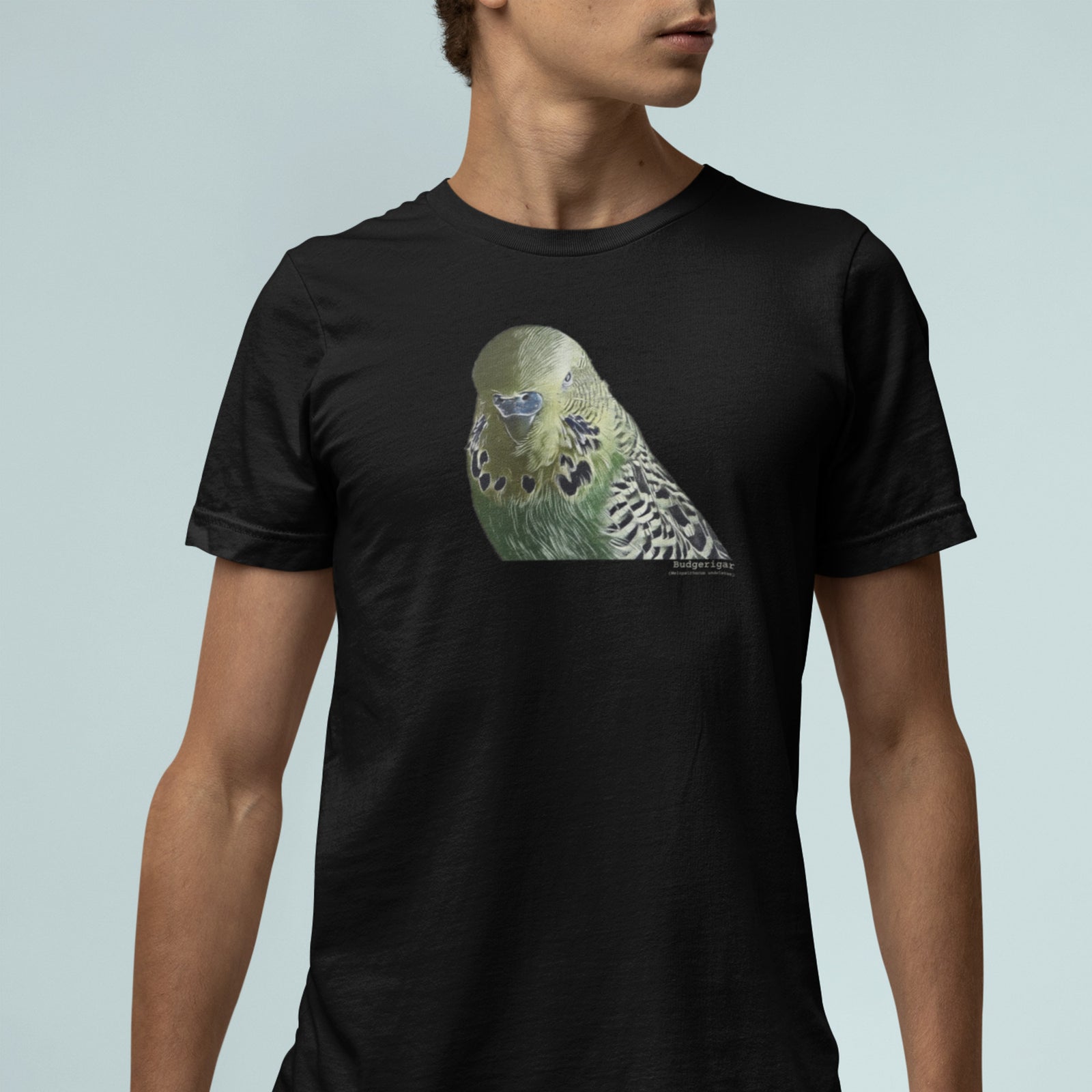 Black unisex t-shirt with an illustrated and lifelike graphic of a green and yellow parakeet or budgie on the front. Below and to the right of the picture is the word Budgerigar and below that the scientific name in parenthesis in small print.