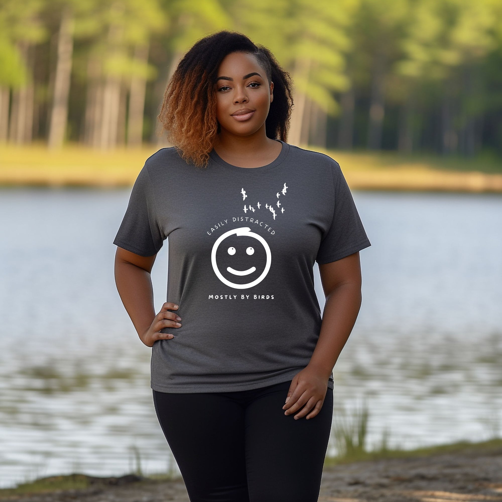 Grey unisex bird t-shirt with a smiley face looking up and to the side at a flock of birds surrounded by the words, "Easily Distracted" above and, "Mostly by Birds" below. Worn by a woman near a river.