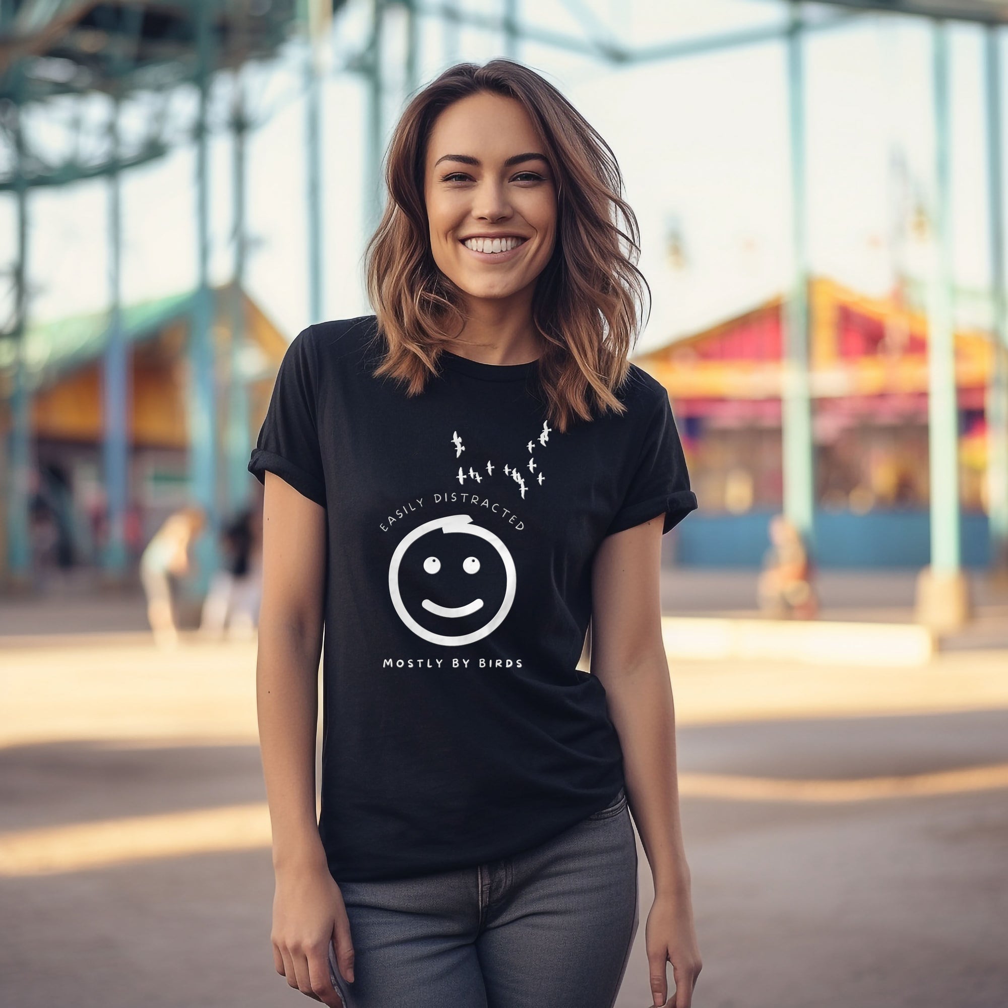 Black unisex bird t-shirt with a smiley face looking up and to the side at a flock of birds surrounded by the words, "Easily Distracted" above and, "Mostly by Birds" below. Worn by a woman at a fairground.