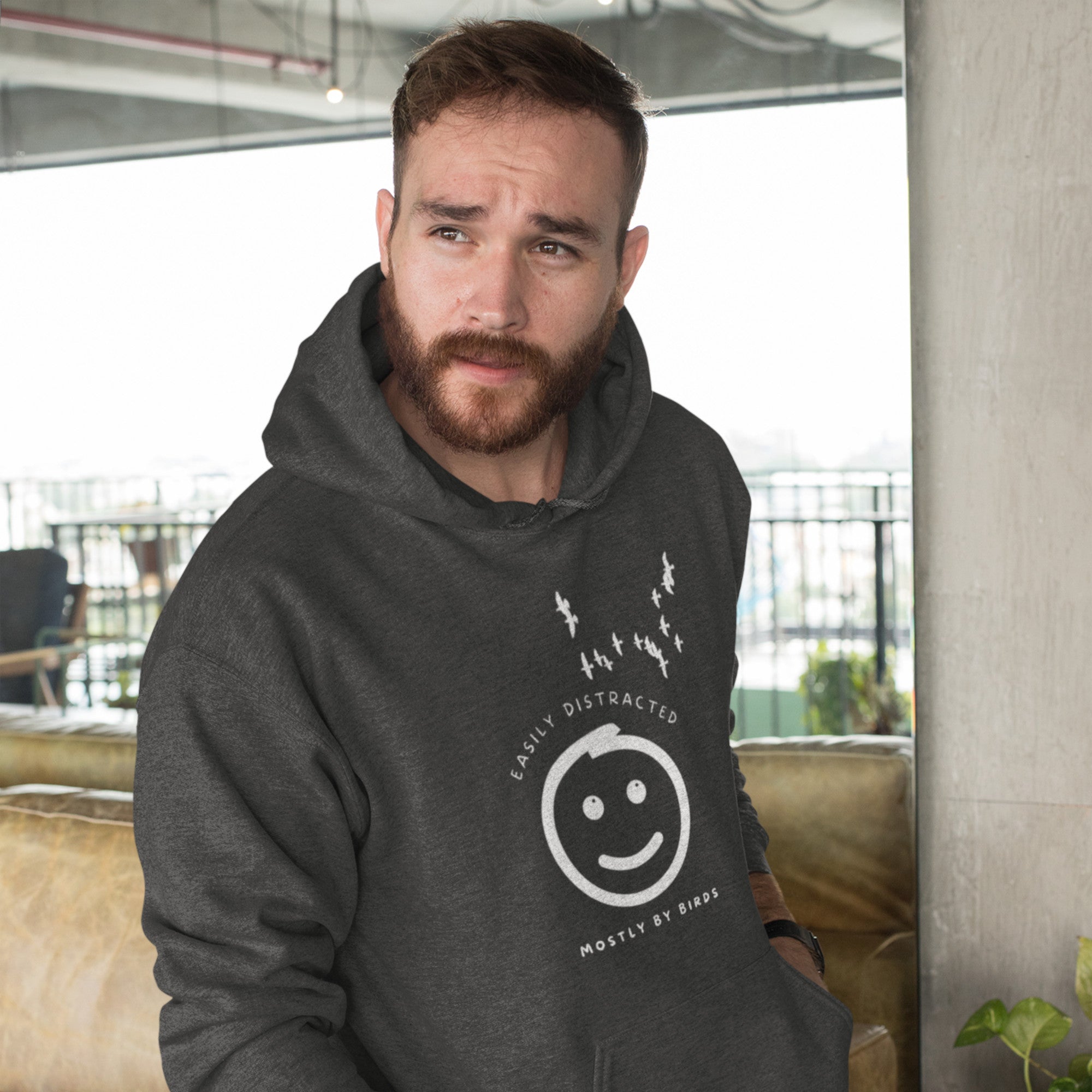 Heather gray hoodie with a smiley face looking off to the right at a flock of birds and the words Easily Distracted, Mostly By Birds on the front. Worn by a man.