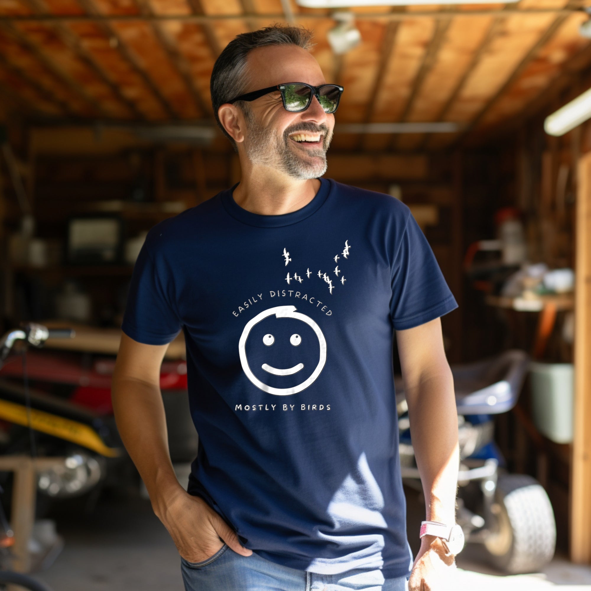 Navy blue unisex bird t-shirt with a smiley face looking up and to the side at a flock of birds surrounded by the words, "Easily Distracted" above and, "Mostly by Birds" below. Worn by a man in a shed or garage.
