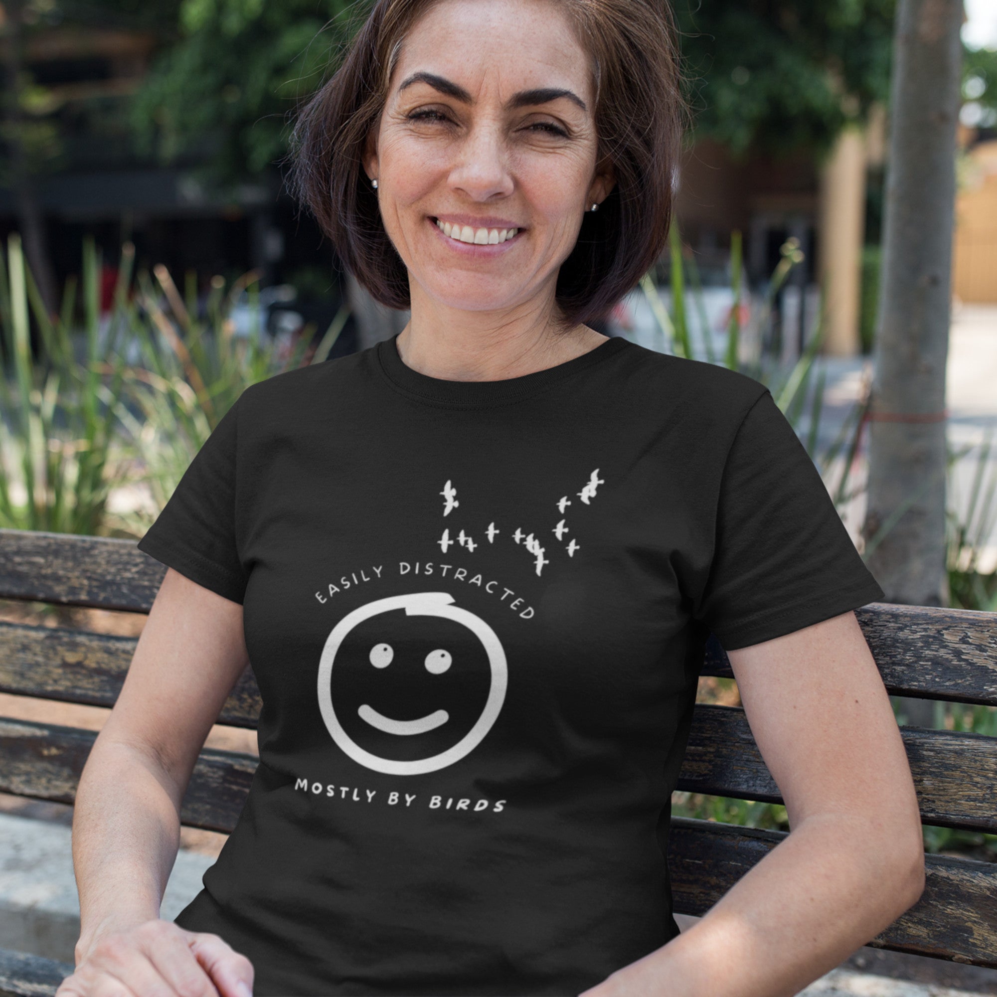 Black unisex bird t-shirt with a smiley face looking up and to the side at a flock of birds surrounded by the words, "Easily Distracted" above and, "Mostly by Birds" below. Worn by a woman sitting on a park bench.