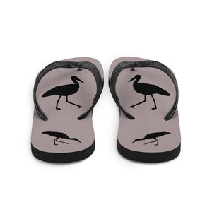 A pair of dusty purple flip-flops with black strap and a silhouette of a stork standing and smaller ones at the heel, viewed from the back.
