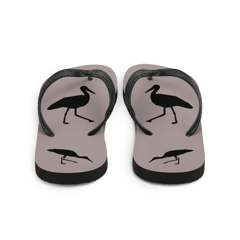 A pair of dusty purple flip-flops with black strap and a silhouette of a stork standing and smaller ones at the heel, viewed from the back.