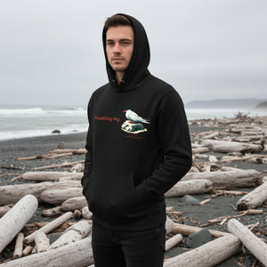 Black unisex hoodie with a graphic of an Arctic Tern on a rock, and the words "Awaiting My" next to it in a fun pun. The words Arctic Tern and the scientific name of Sterna Paradisaea are below the bird. Worn by a man standing on a rocky, driftwood laden beach.