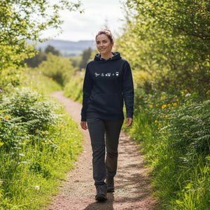 Unisex navy blue hooded bird sweatshirt celebrating bird watching and coffee with a graphic of binoculars, a bird, and a travel cup of coffee. Worn by a woman walking a nature trail.