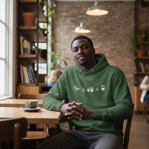Unisex forest green hooded bird sweatshirt celebrating bird watching and coffee with a graphic of binoculars, a bird, and a travel cup of coffee. Worn by a man in a coffee shop.