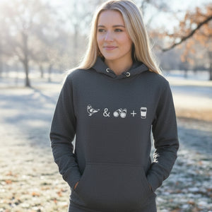 Unisex dark heather grey hooded bird sweatshirt celebrating bird watching and coffee with a graphic of binoculars, a bird, and a travel cup of coffee. Worn by a woman in a park on a cold day.