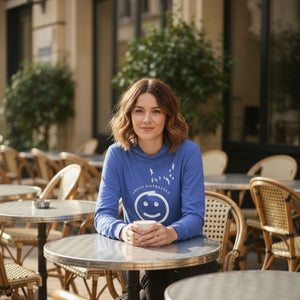 Heather royal blue long sleeved and hooded tee with fun, "Easily Distracted, Mostly by Birds" saying on the front, with a smiley face looking up at a flock of birds. Worn by a woman sitting at an outdoor cafe.