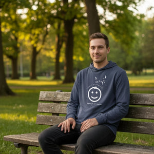 Heather navy blue long sleeved and hooded tee with fun, "Easily Distracted, Mostly by Birds" saying on the front, with a smiley face looking up at a flock of birds. Worn by a man sitting in a park.