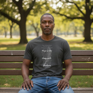 Man sitting on a bench in a park wearing a t-shirt with Emily Dickinson's Hope is the Thing with Feathers poem in white text.