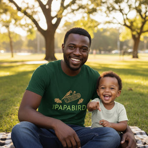 Man and child sitting in a park, the man is wearing an emerald green 'The PAPABIRD' t-shirt featuring a woodpecker on a log and oak leaves.