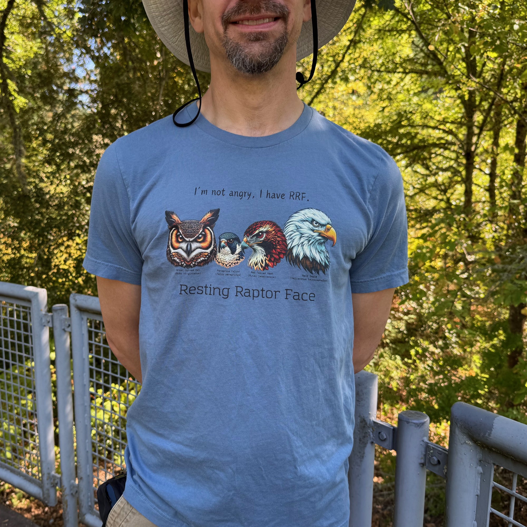 A steel blue bird shirt with a great horned owl, a peregrine falcon, a red-tailed hawk and a bald eagle. The shirt has text that says, I'm not angry, I have RRF. Resting Raptor Face. Worn by a man.