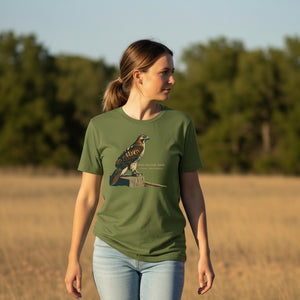 Green bird shirt with an artistic depiction of a red-tailed hawk sitting on a fence and the common and scientific names for the bird next to it. Worn by a woman walking in a field.
