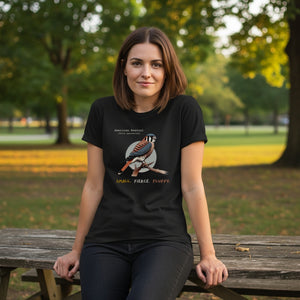 A black women's bird shirt with the words, "Small. Fierce. Fluffy" below an illustration of an American Kestrel. Worn by a woman sitting on a picnic table.