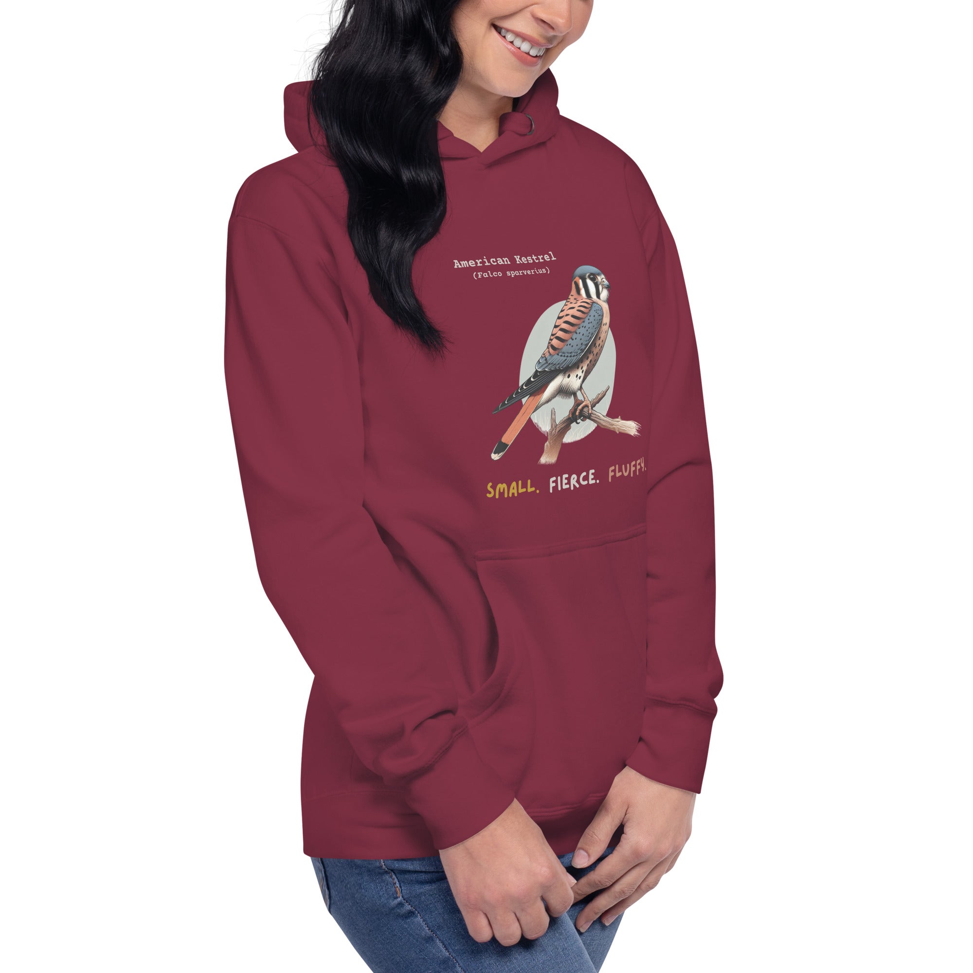 Maroon  hooded unisex bird sweatshirt with the words, "Small. Fierce. Fluffy" below an illustration of an American Kestrel. Worn by a woman.