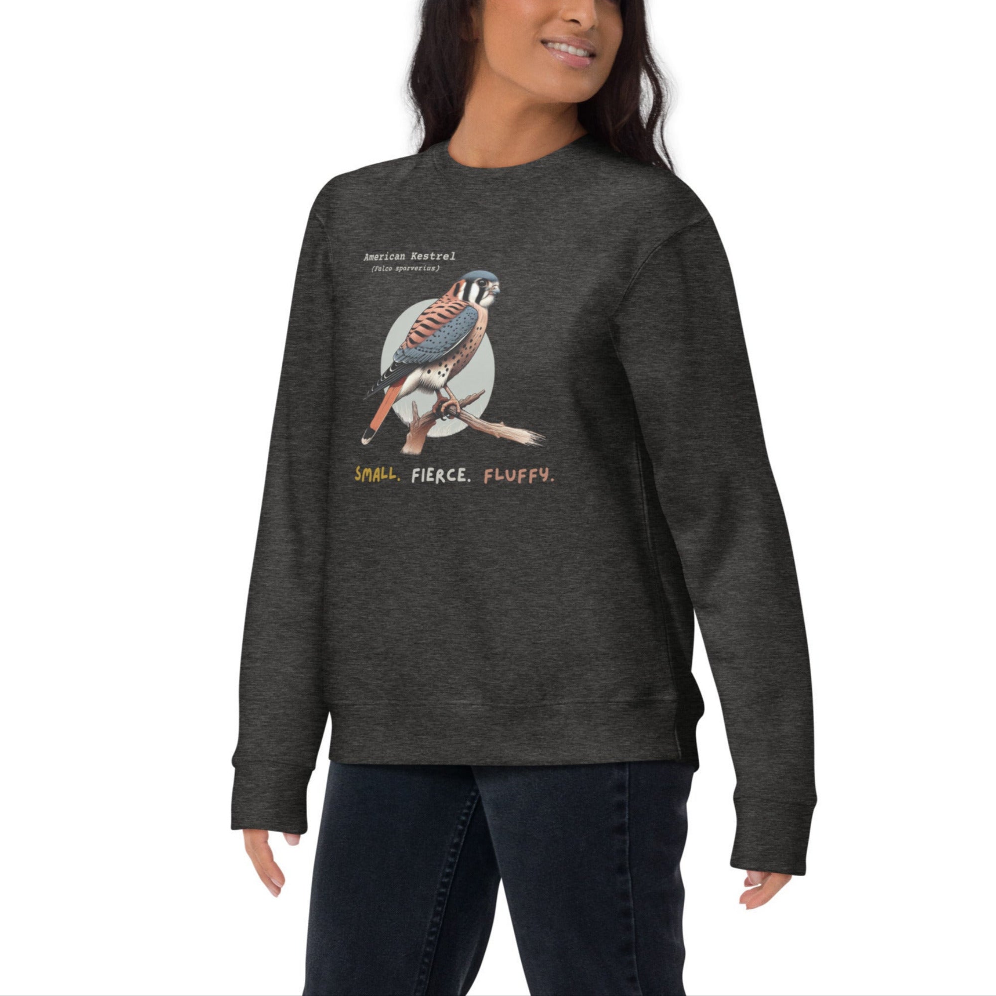 Dark heather gray unisex bird sweatshirt with the words, "Small. Fierce. Fluffy" below an illustration of an American Kestrel. Worn by a woman.