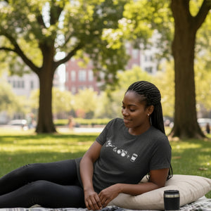 Dark heather gray women's bird t-shirt celebrating bird watching and coffee with a graphic of binoculars, a bird, and a travel cup of coffee. Worn by a woman relaxing on a blanket in a park.